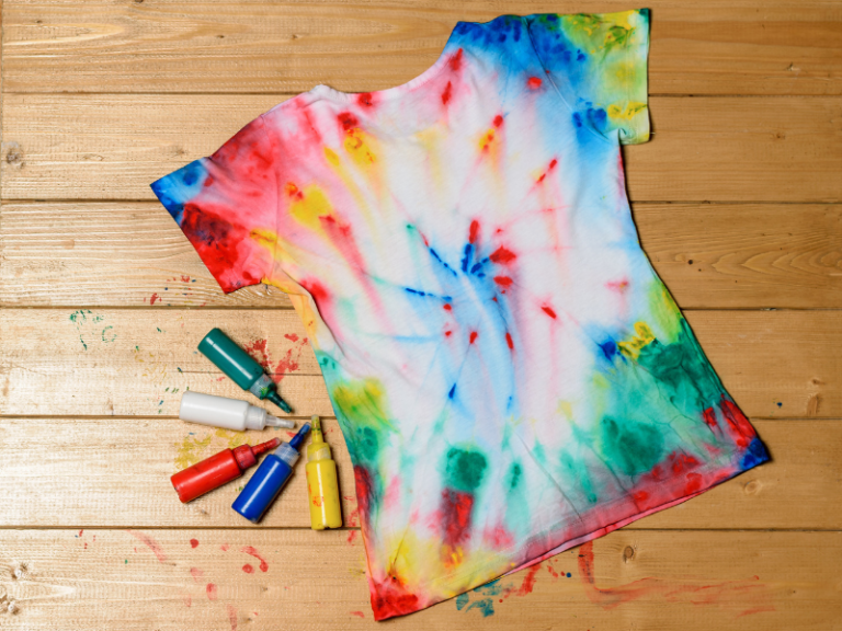 tie-dyed t-shirt on a wooden floor with colors paints and paint splatter next to it.